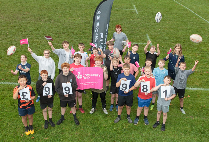 Group of teenagers smiling holding A4 numbers and National Lottery branding some throwing rugby balls in the air with Strathmore Community Rugby Trust feather flag behind